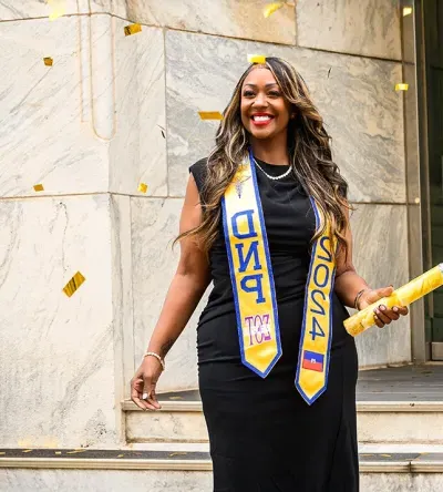 Graduating student celebrating with confetti, wearing a black dress and DNP 2024 sash, holding a diploma outside a building.